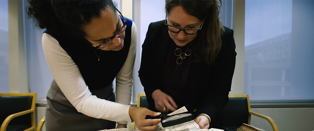 TWO WOMEN USE MAGNIFYING GLASS TO EXAMINE DOCUMENT